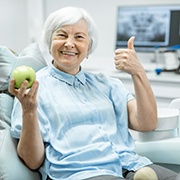 An older woman holding an apple and smiling after receiving dental implants in Oklahoma City